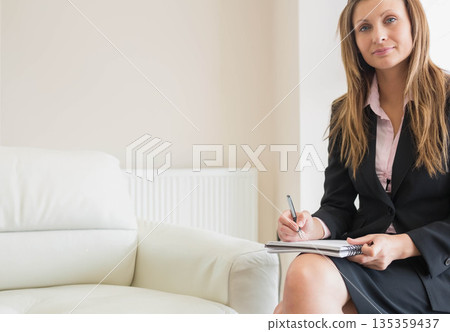Executive woman sitting on leather sofa in office holding notebook and pen poised, copy space Executive woman sitting on leather sofa in office holding notebook and pen poised, copy space 135359437