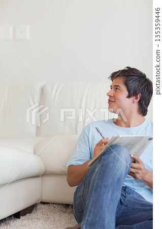 Man sitting in living room leaning against leather sofa holding pen and newspaper, copy space 135359446