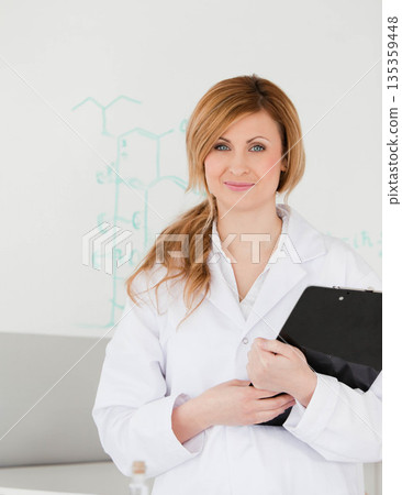 Female scientist wearing lab coat standing in lab by whiteboard and holding clipboard at counter 135359448