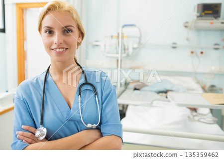 Adult female nurse wearing scrubs and stethoscope standing by bed in hospital room, copy space 135359462