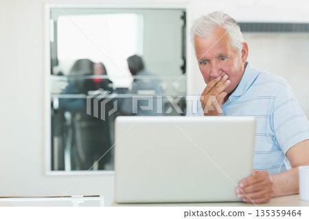 Senior man reacting with surprise at office desk examining laptop and coffee mug by glass partition 135359464