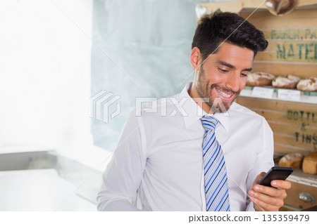 Man wearing business attire checking smartphone at bakery with wooden bread shelves and chalkboard Man wearing business attire checking smartphone at bakery with wooden bread shelves and chalkboard 135359479