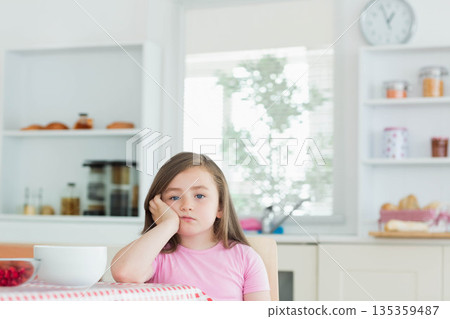 Child girl resting cheek on hand and gazing at kitchen table with berry bowls, checkered tablecloth 135359487