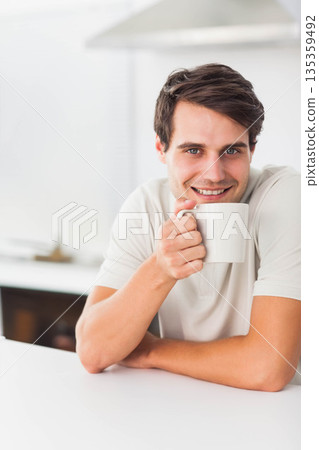Man in his twenties sitting at kitchen counter holding white mug near mouth, smiling, copy space 135359492
