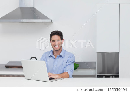 Man sitting at white countertop in modern kitchen working on silver laptop near green plant Man sitting at white countertop in modern kitchen working on silver laptop near green plant 135359494