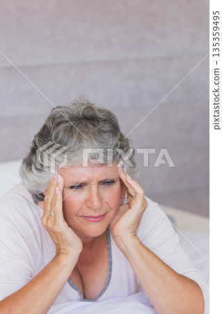Senior woman leaning forward on bed pressing temples with pillow by pastel wall in pink sleepwear 135359495