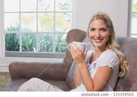 Woman sitting on sofa by multi-pane window holding white ceramic mug near chest, copy space Woman sitting on sofa by multi-pane window holding white ceramic mug near chest, copy space 135359503