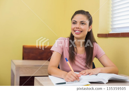 Female student sitting at wooden desk in study room holding pencil and writing from open textbook 135359505