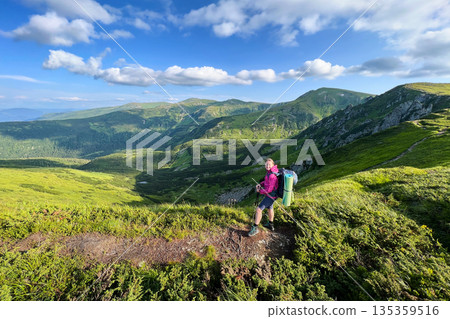 Woman hiker stands on mountain trail, taking in breathtaking view of rolling green hills and valleys. Backpack and sleeping pad visible as she grips trekking poles under partly cloudy sky. Woman hiker stands on mountain trail, taking in breathtaking view of rolling green hills and valleys. Backpack and sleeping pad visible as she grips trekking poles under partly cloudy sky. 135359516