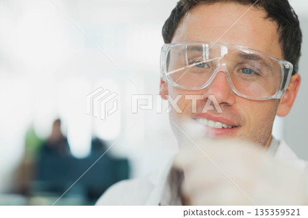Man examining pipette under bright lighting in lab wearing lab coat and goggles, copy space 135359521