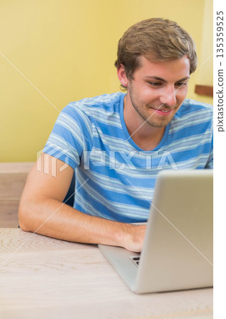 Man leaning forward while typing on silver laptop at light woodgrain table inside pale yellow room Man leaning forward while typing on silver laptop at light woodgrain table inside pale yellow room 135359525