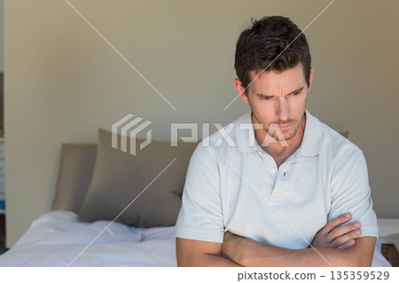 Man wearing white polo sitting on bed edge gazing in bedroom with taupe pillows, copy space Man wearing white polo sitting on bed edge gazing in bedroom with taupe pillows, copy space 135359529