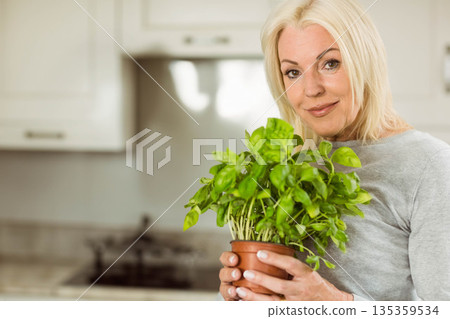 Senior woman holding pot with bright green herb in home kitchen with white cabinets, copy space 135359534