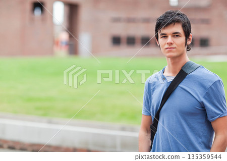 Male student standing on grass lawn near brick campus building with bag strap, copy space Male student standing on grass lawn near brick campus building with bag strap, copy space 135359544