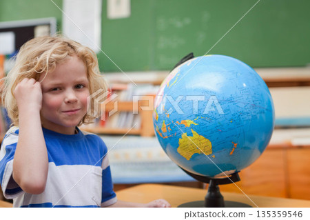 Blonde boy sitting at desk in classroom, raising hand while studying globe on stand, copy space Blonde boy sitting at desk in classroom, raising hand while studying globe on stand, copy space 135359546