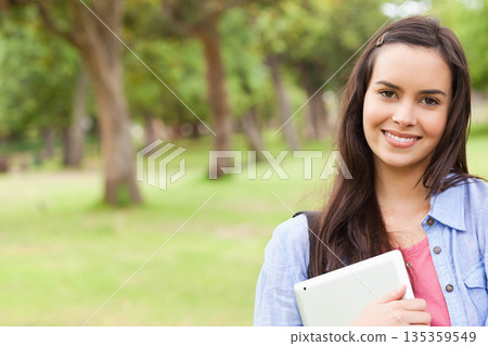 Female student standing in park holding white tablet and smiling with backpack strap, copy space Female student standing in park holding white tablet and smiling with backpack strap, copy space 135359549