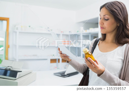 Woman holding amber medicine bottle and info sheet at pharmacy counter using tablet, copy space 135359557