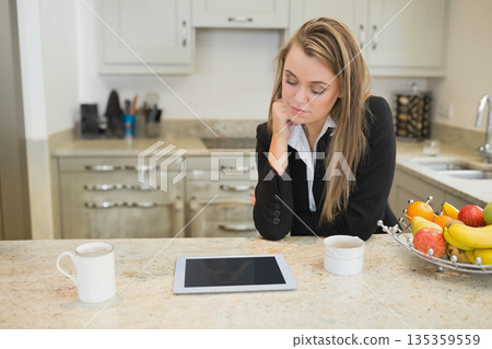 Businesswoman leaning on one hand at kitchen counter studying tablet between mugs and fruit bowl 135359559