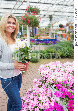 Woman holding red pot of white flowers while standing on brick aisle in greenhouse, copy space Woman holding red pot of white flowers while standing on brick aisle in greenhouse, copy space 135359565