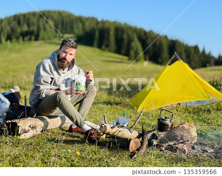 Man tourist eating from food pouch with green spoon, sits on log near campfire. Yellow tent on green hillside, surrounded by lush trees. Peacefulness of camping in beautiful natural environment. Man tourist eating from food pouch with green spoon, sits on log near campfire. Yellow tent on green hillside, surrounded by lush trees. Peacefulness of camping in beautiful natural environment. 135359566