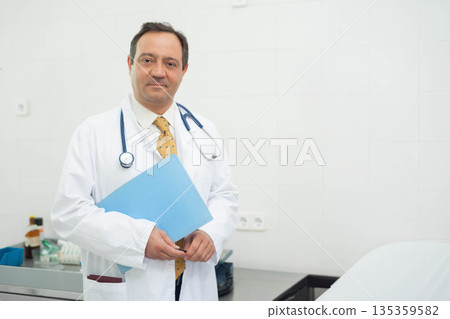 Middle-aged physician holding blue folder in exam room with stethoscope and lab coat, copy space 135359582