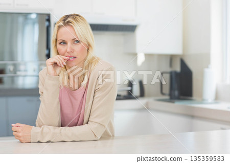 Middle-aged woman leaning forward touching lips and gazing left at kitchen countertop, copy space 135359583