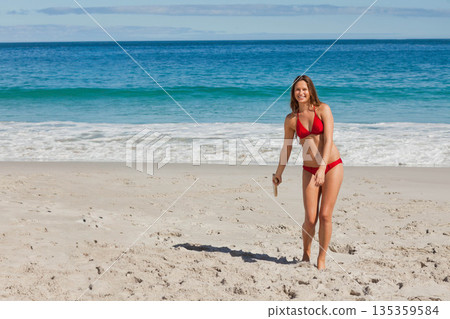 Woman standing on sand near water edge wearing red bikini holding wooden boomerang, copy space 135359584