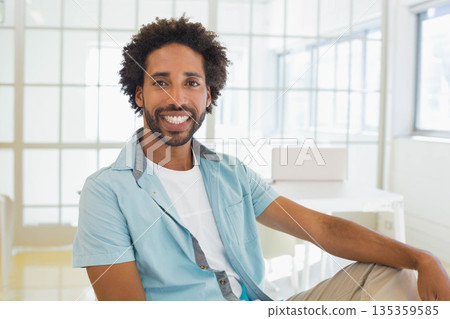 African American man wearing light blue shirt sitting casually in office with glass panels and desk African American man wearing light blue shirt sitting casually in office with glass panels and desk 135359585