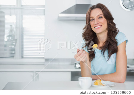 Woman holding fork lifting cake bite while smiling at kitchen counter with mug, copy space 135359600