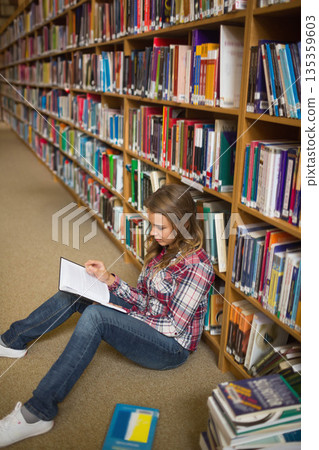 Woman sitting on carpeted library aisle holding open hardcover book with stack of books, copy space 135359603