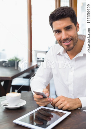 Male in 30s wearing white shirt sitting at cafe with smartphone, tablet, cappuccino, copy space 135359609