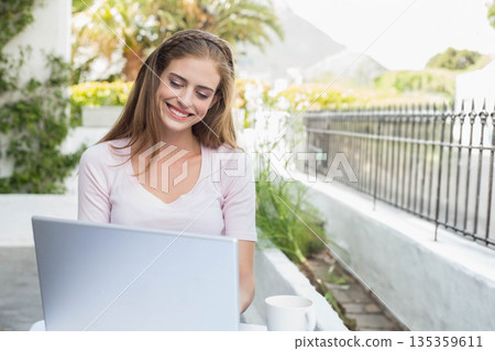 Woman working on silver laptop at white patio table on bright terrace, holding coffee mug 135359611