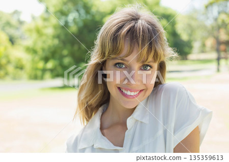 Smiling woman standing outdoors in sunlit park highlighting green eyes and collared blouse Smiling woman standing outdoors in sunlit park highlighting green eyes and collared blouse 135359613