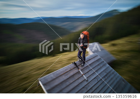 Hiker with orange backpack stands on roof of wooden cabin, surrounded by blurred, dynamic scenery. Motion blur effect. 135359616