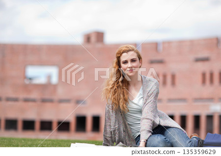 Female student sitting on lawn in front of brick building studying with open notebooks, copy space 135359629