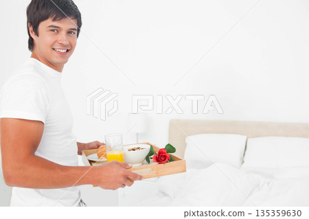 Man in twenties carrying tray with orange juice croissant rose to bed in bedroom, copy space Man in twenties carrying tray with orange juice croissant rose to bed in bedroom, copy space 135359630
