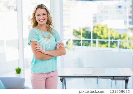 Smiling woman standing arms crossed in office wearing mint top near table by windows, copy space 135359635