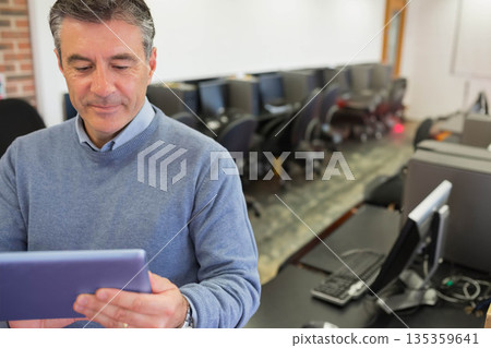 Senior man in light blue sweater standing in computer lab holding tablet near monitors, copy space Senior man in light blue sweater standing in computer lab holding tablet near monitors, copy space 135359641