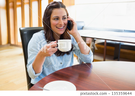 Woman in denim shirt sitting at cafe table holding white ceramic mug and talking on smartphone Woman in denim shirt sitting at cafe table holding white ceramic mug and talking on smartphone 135359644