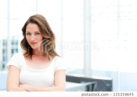 Mid adult woman standing with arms crossed in office by large windows with blinds, copy space 135359645
