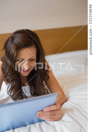 Female reclining on bed in bedroom holding blue tablet and smiling and wearing white nightwear 135359646