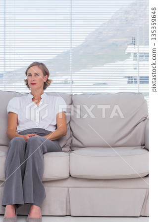 Middle-aged woman sitting on beige sofa at home wearing blouse, trousers, facing blinds, copy space 135359648