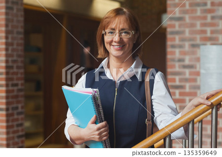 Middle-aged woman standing on staircase in brick library holding notebooks wearing shoulder bag Middle-aged woman standing on staircase in brick library holding notebooks wearing shoulder bag 135359649