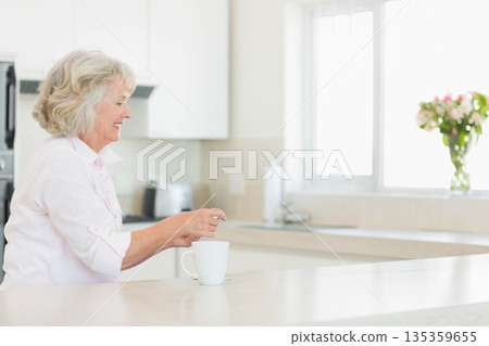 Senior woman standing at kitchen counter stirring white mug beside toaster and vase of flowers Senior woman standing at kitchen counter stirring white mug beside toaster and vase of flowers 135359655
