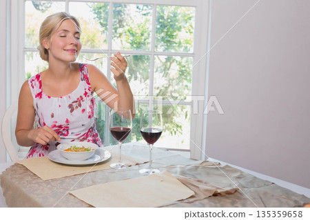 Woman in twenties eating salad from bowl at dining table near window with wine, copy space 135359658