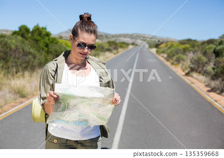 Middle-aged woman standing on rural road holding paper map wearing backpack with mat, sunglasses 135359668