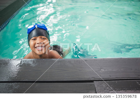 African American boy emerging from pool, leaning on wood deck in black cap and blue goggles African American boy emerging from pool, leaning on wood deck in black cap and blue goggles 135359678