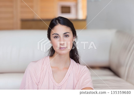 Light-skinned woman sitting on pale couch in living room with wood shelving and cabinet 135359680