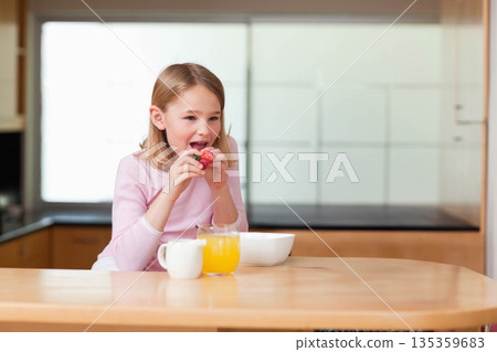 Female child biting strawberry at wooden kitchen counter with bowl, juice and mug, copy space 135359683