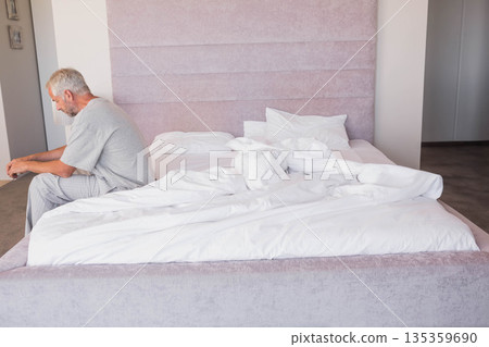 Senior man sitting on edge of unmade bed in pastel-toned bedroom with padded headboard, copy space 135359690